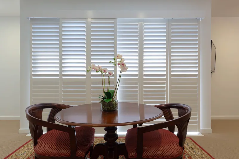 Round dining table in front of full-height plantation shutters