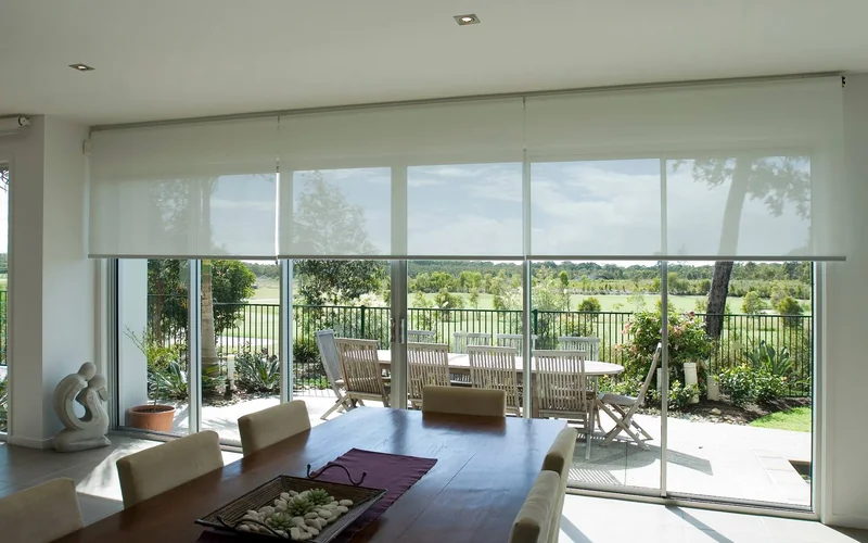 Sunscreen roller blinds drawn across sliding doors in a Brisbane dining room overlooking a garden